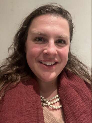 A woman with long brown hair smiles warmly at the camera. She wears a maroon scarf, a beige top, and a layered white bead necklace. Neutral background.