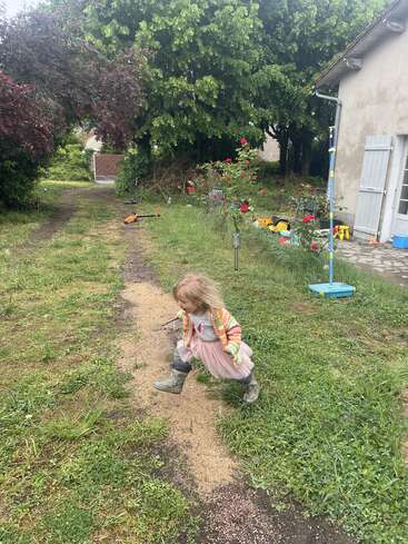 A young girl in a pink dress and boots is running outside a house, surrounded by grass, trees, flowers, and colorful outdoor toys. It looks playful.