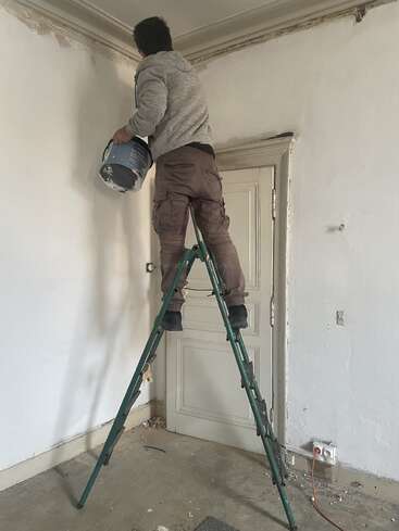 A person stands on a green ladder, holding a paint bucket, preparing to paint or repair the upper corner of a white, partially renovated room.