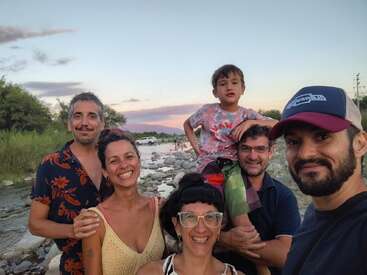 Six smiling adults and one child pose together outdoors by a rocky river at sunset, surrounded by greenery. The mood is cheerful and relaxed, enjoying nature.