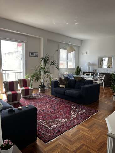 Un salon lumineux avec de grandes fenêtres, des canapés bleu marine, des fauteuils rayés, un tapis à motifs rouges, un sol en bois, des plantes vertes, une table de salle à manger blanche et des accessoires décoratifs créent une atmosphère chaleureuse.