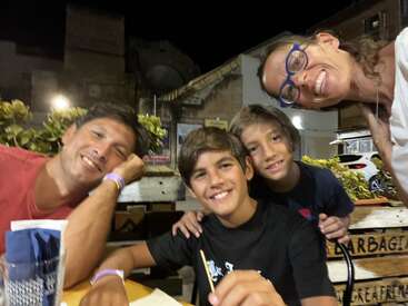 Uma família feliz de quatro pessoas posa junta, sorrindo em um restaurante ao ar livre à noite. Há plantas atrás deles e a iluminação ambiente quente cria uma atmosfera aconchegante.