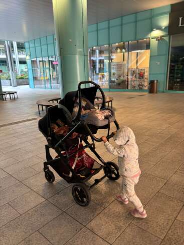 A small child in a hooded outfit pushes a stroller with a baby inside through a modern, covered shopping area with glass walls and tiled floor.