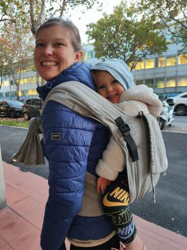 A smiling woman in a blue jacket carries a happy child on her back. They are outside near parked cars and trees, both warmly dressed.