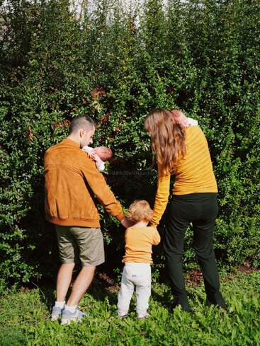 A family of four stands outdoors, facing greenery. The parents each hold a baby, while their toddler stands between them, holding their hands. Sunny day.