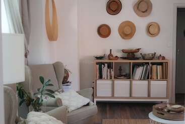 Un salon confortable avec des fauteuils, une étagère en bois contenant des livres et des objets de décoration, des plantes en pot et des chapeaux artistiquement disposés sur le mur blanc pour la décoration.