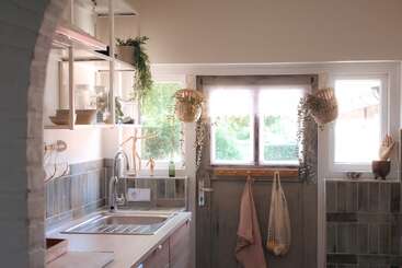 Une cuisine confortable avec lumière naturelle, armoires en bois, carreaux verts, paniers suspendus, plantes, torchons et bols en céramique. Atmosphère calme, décor accueillant et chaleureux, charme rustique.