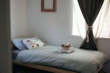 Une petite chambre confortable avec un lit simple, une literie légère, un oreiller fleuri, des serviettes pliées, un panier, une grande fenêtre avec des rideaux attachés et un cadre photo.