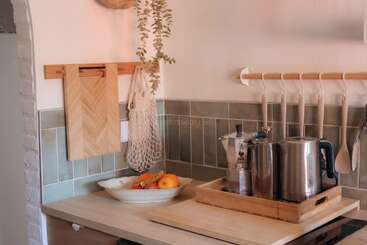 Un coin cuisine confortable avec des plans de travail en bois, des planches à découper, des ustensiles suspendus, un plateau avec une bouilloire et une cafetière, une coupe de fruits et des carreaux muraux verts.