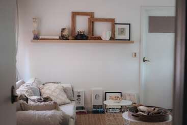 Un salon confortable avec un canapé blanc, des coussins neutres, des cadres décoratifs et des œuvres d'art sur une étagère flottante. Deux tables d'appoint accueillent des bols et des couvertures.