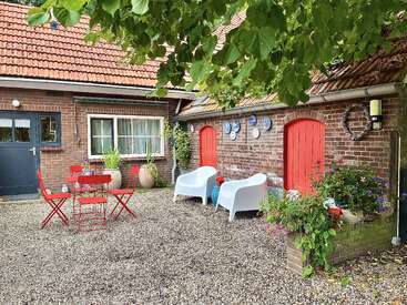 Une charmante cour avec des murs en briques rouges, des portes rouges, des chaises blanches, une table rouge, des plantes en pot, des assiettes décoratives, un sol en gravier et des branches d'arbre vertes en surplomb.