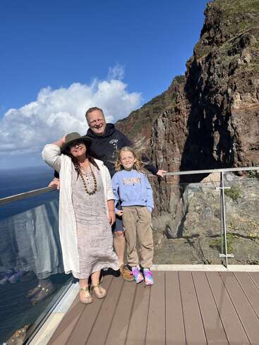 Une famille de trois personnes pose sur une vue panoramique de l'océan et des falaises. La journée est ensoleillée, le ciel est bleu, les nuages sont cotonneux et tout le monde a l'air heureux.