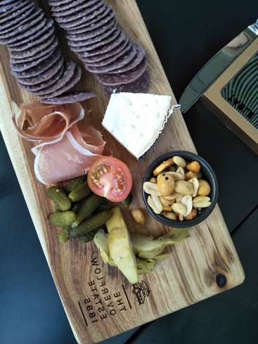 A imagem mostra uma tábua de charcutaria com biscoitos, queijo, carne, nozes, picles e legumes em uma tábua de madeira com um logotipo onde se lê \"THE OVERFLOW ESTATE 1885\".