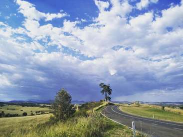 A imagem retrata uma paisagem rural serena com uma estrada sinuosa, árvores e colinas ondulantes sob um céu azul com nuvens brancas.