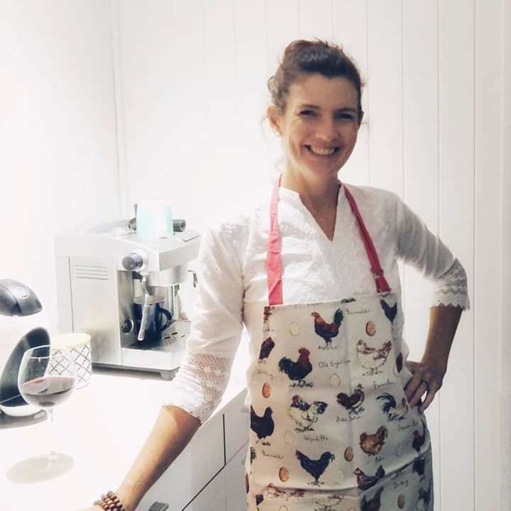 A imagem mostra uma mulher em pé em uma cozinha, usando um avental com desenhos de galinhas, com uma máquina de café e uma taça de vinho no balcão atrás dela.