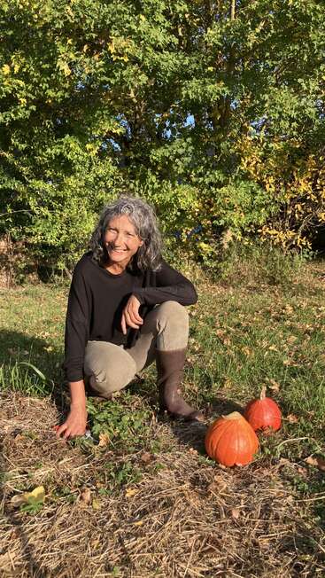 Una mujer de pelo rizado y gris sonríe arrodillada al aire libre. Está cerca de tres calabazas en el suelo, rodeada de follaje verde y árboles.