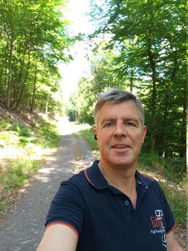 Um homem tira uma selfie em um dia ensolarado enquanto caminha em uma trilha de cascalho em uma floresta verde e exuberante. Ele está sorrindo e vestindo uma camisa polo azul-marinho.