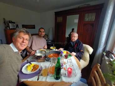 Três homens estão sentados ao redor de uma mesa de jantar, saboreando uma refeição caseira, com bebidas, comida e uma vela acesa, criando uma atmosfera calorosa e aconchegante.