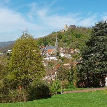 Um vilarejo pitoresco aninha-se entre colinas verdes, dominado por um castelo no topo da colina. Uma igreja com uma alta torre fica entre casas charmosas, sob um céu azul brilhante.