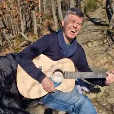 A man is joyfully playing an acoustic guitar outdoors. He is sitting on a wooden bench, surrounded by trees, enjoying the sunlight and nature.