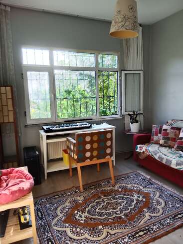 Une pièce confortable avec une grande fenêtre, un tapis à motifs, une chaise en patchwork colorée, un canapé rouge, un clavier sur un bureau blanc, une plante d'intérieur et une lumière naturelle chaude.