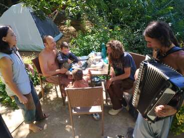 Five adults and two children gather around a table outdoors, enjoying food together beside a tent. One person is playing an accordion, surrounded by lush greenery.