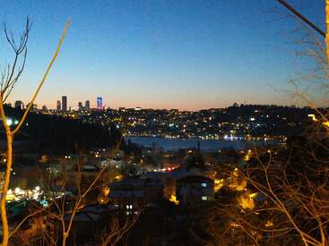 This image captures a stunning cityscape at dusk, with glowing city lights, high-rise buildings, a calm river, silhouetted trees, and a brilliant blue-orange sky.