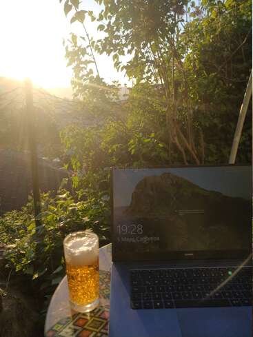 A sunny outdoor setting features a glass of sparkling drink, a patterned table, and a laptop displaying the time 19:28, surrounded by lush greenery.