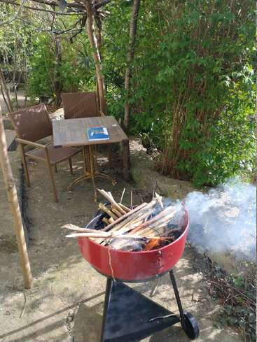 A red barbecue grill with smoking wood sits on a patio. Nearby, two chairs and a table with a book offer a peaceful, green garden setting.