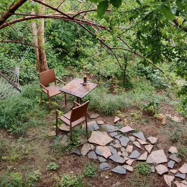 Two brown chairs and a small wooden table sit on a stone path in a lush green garden. A hammock hangs nearby among the trees.