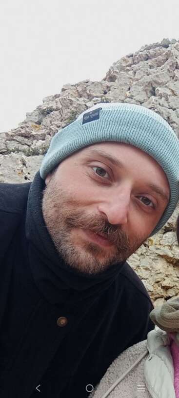 A man with a beard and mustache wears a light blue beanie and black jacket, standing outdoors near rocky terrain, smiling gently at the camera.