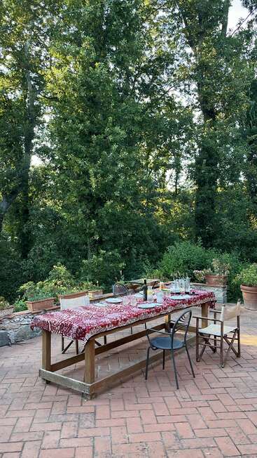 An outdoor dining table sits on a brick patio, covered in a red patterned cloth, surrounded by chairs, wine, glasses, dishes, and lush green trees.
