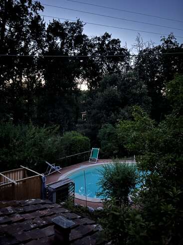 A serene backyard scene at dusk, featuring a tranquil swimming pool surrounded by lush greenery, lounge chairs, and dense trees silhouetted against the fading evening sky.