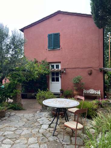 A charming pink house with green shutters, surrounded by lush greenery, a stone patio, round table, striped chairs, white bench, and vibrant potted plants throughout.