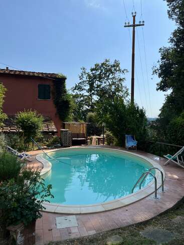 A serene backyard with a round swimming pool, surrounded by lush greenery, deck chairs, a rustic house with green shutters, and a tall power line pole.