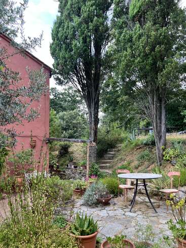 A charming garden patio with potted plants, stone steps leading uphill, two pink chairs, a round table, tall cypress trees, and a rustic red house exterior.