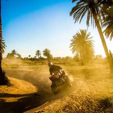 La imagen muestra a dos individuos conduciendo un quad por un camino de tierra, uno en primer plano y el otro al fondo, en un entorno natural al aire libre.