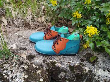 Um par de sapatos de couro turquesa e marrom com cadarços escuros está apoiado sobre uma superfície de pedra ao ar livre, rodeado por plantas verdes e flores amarelas.
