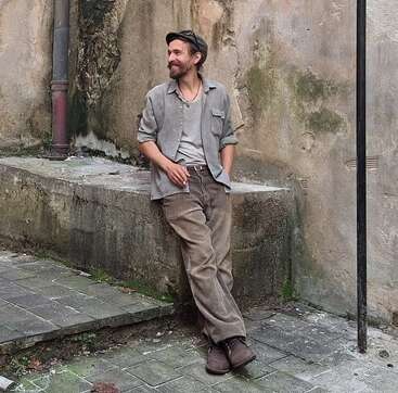 Um homem descontraído, vestindo roupas casuais de estilo vintage, está encostado em um parapeito de pedra numa rua antiga, sorrindo, ao lado de uma parede desgastada pelo tempo e coberta de musgo.