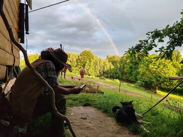 Una persona está sentada junto a una cabaña de madera hablando por teléfono, mientras un perro descansa a su lado. Los caballos pastan bajo los árboles y un hermoso arco iris se extiende por el cielo.