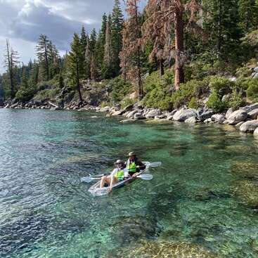 A man and woman paddle a clear kayak through a serene lake, surrounded by large rocks and trees, with a cloudy sky above.