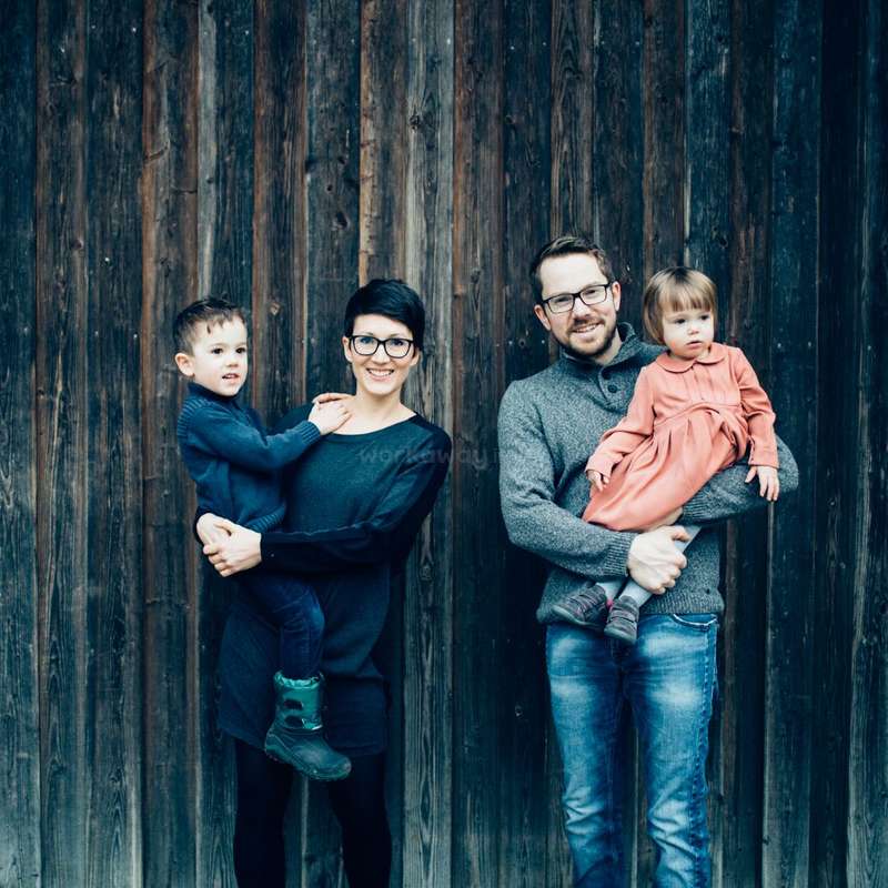 L'image représente une famille de quatre personnes debout devant un mur en bois, les parents tenant leurs deux jeunes enfants. La famille est habillée de manière décontractée.