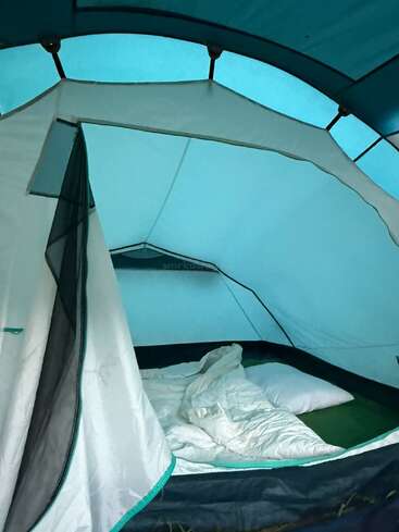 Dentro de uma barraca de acampamento azul, um saco de dormir branco e um travesseiro estão sobre um colchão inflável. A suave luz da manhã penetra pelas janelas de tela, criando um ambiente aconchegante.