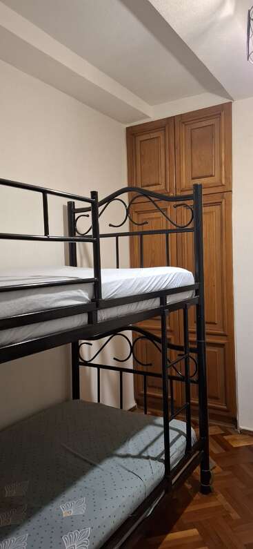 Dieses Bild zeigt ein einfaches Schlafzimmer mit einem Etagenbett aus Metall, einer mit Laken bezogenen Matratze und einem hölzernen Kleiderschrank vor einer cremefarbenen Wand.