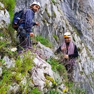 Dos hombres de pie en un acantilado rocoso, con cascos y sujetando cuerdas de escalada, con una mochila y flores silvestres que añaden un toque de aventura al escarpado paisaje.