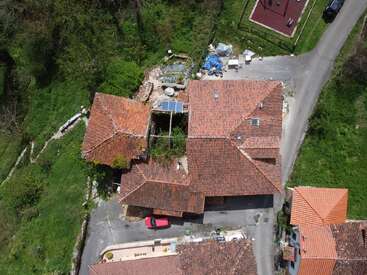 Esta imagem aérea mostra uma casa com telhados de telhas vermelhas, cercada por vegetação e ruas. Dá pra ver carros, painéis solares, áreas ajardinadas e vários objetos ao ar livre.