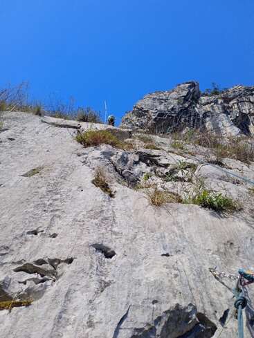 Uma falésia rochosa íngreme sob um céu azul vibrante exibe manchas de grama, equipamentos de escalada à vista e um alpinista no topo, destacando uma escalada cheia de aventura.