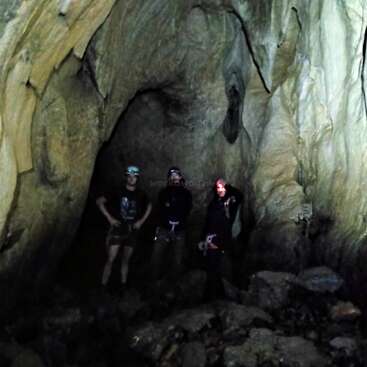 Três pessoas com lanternas de cabeça estão dentro de uma grande caverna rochosa. A luz fraca destaca as paredes irregulares da caverna e o chão acidentado, coberto de pedras e sombras.