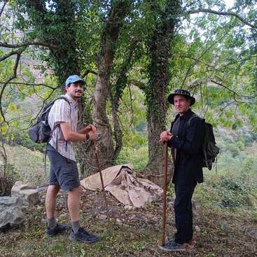 Dois homens com mochilas e bastões de caminhada estão em uma trilha numa área florestal. Eles estão sorrindo, rodeados por árvores, com uma lona atrás deles.