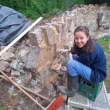 Eine Frau in Handschuhen und Stiefeln restauriert draußen fröhlich eine Steinmauer. Umgeben von Werkzeugen und Grünpflanzen hockt sie auf einer Kiste und arbeitet mit einer Kelle.